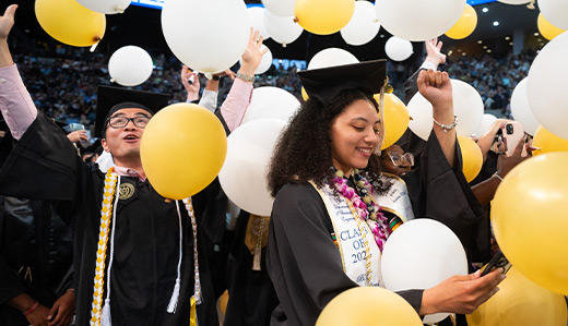 Georgia Tech students wearing full regalia smile and celebrate at commencement. White and gold ballons are filling the frame as they are dropped from the ceiling